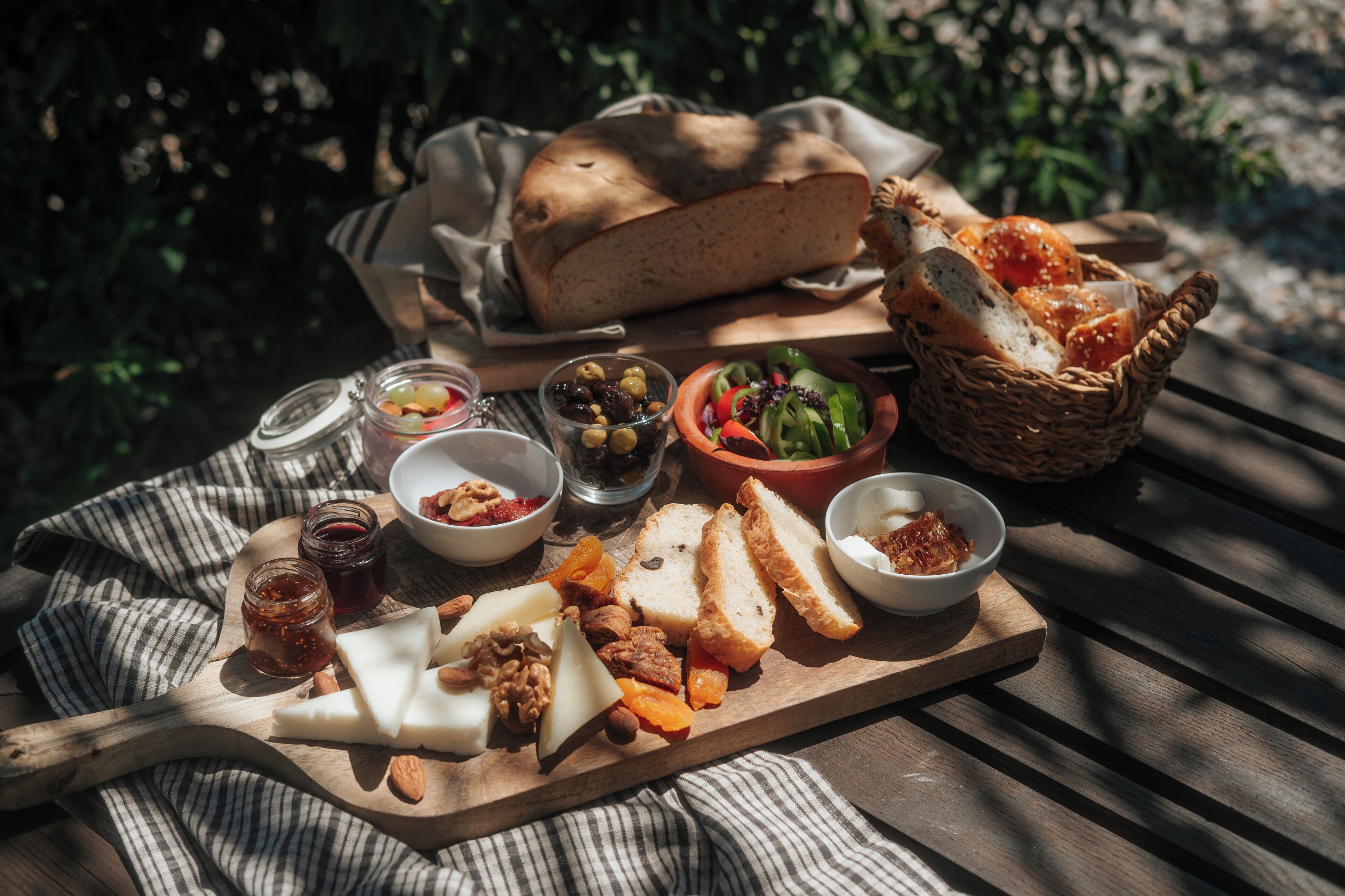 A picnic setup with bread, cheese, nuts, and various spreads on a wooden board, set outdoors.