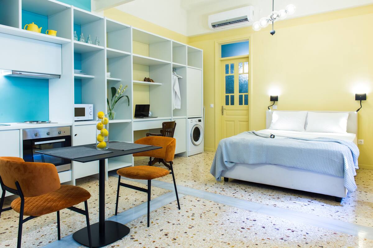 A compact room with a bed, a dining table with chairs, a washing machine, and a kitchenette.