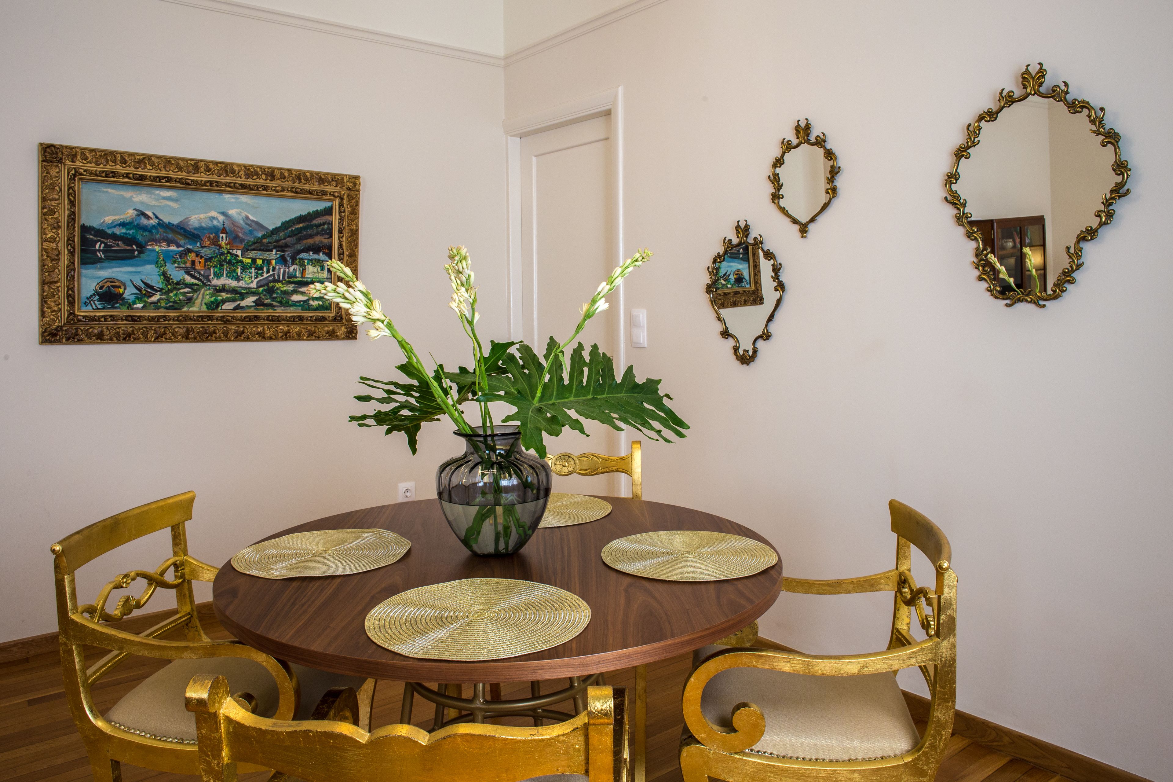 A dining area with a round wooden table, gold chairs, and a vase with green plants.