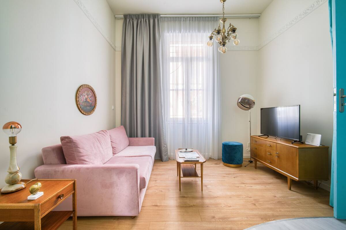 A living room with a pink sofa, a wooden coffee table, a TV on a wooden cabinet, and a large window with sheer curtains.