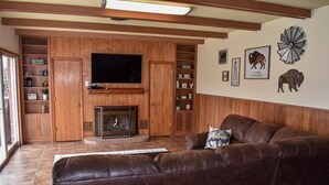 A living room with a brown leather sofa, a flat-screen TV, wooden shelves, and a fireplace. - The Cozy Buffalo near Palo Duro Canyon & WTAMU (Canyon)