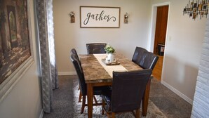 A dining area with a wooden table, black leather chairs, a decorative centerpiece, and a 'gather' sign on the wall. - The Cozy Buffalo near Palo Duro Canyon & WTAMU (Canyon)