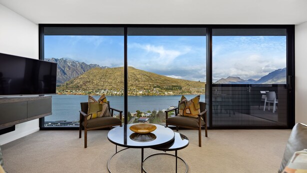Een moderne woonkamer met een grote televisie, twee stoelen, een klein tafeltje en uitzicht op een meer en bergen door grote ramen.