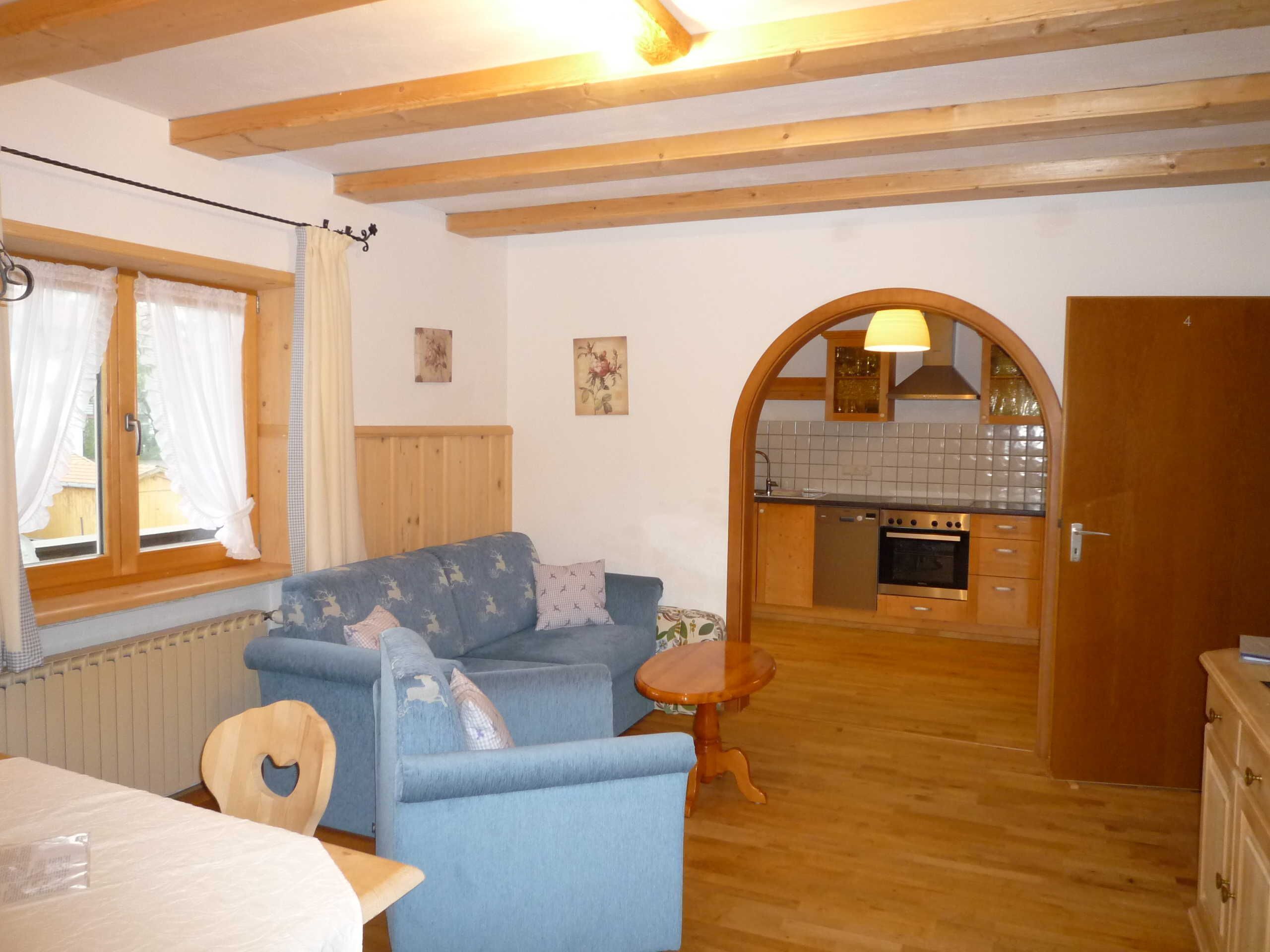 Ein gemütliches Zimmer mit einer Couch, einem kleinen Holztisch, einer Küchenzeile mit Herd und Schränken sowie einem Fenster mit Vorhängen.