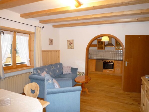 A cozy room with a sofa, a small wooden table, a kitchen area with a stove and cabinets, and a window with curtains. - Apartment 1st floor left - Guesthouse Johanna (Bad Wiessee)