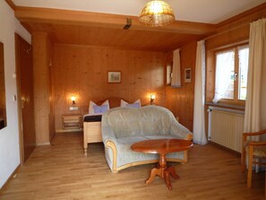 A wooden-paneled room with a bed, a sofa, a round wooden table, and a window with curtains. - Apartment 1st floor left - Guesthouse Johanna (Bad Wiessee)