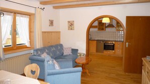 Una habitación acogedora con sofá, una pequeña mesa de madera, una zona de cocina con estufa y armarios, y una ventana con cortinas.