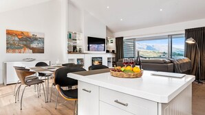 Une cuisine moderne dotée d’un îlot central, d’un coin repas avec des chaises, d’un téléviseur à écran plat et d’une grande fenêtre offrant une vue sur les montagnes.