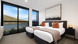 A modern bedroom with two beds, a large window with a view of a lake and hills, and a minimalist decor.