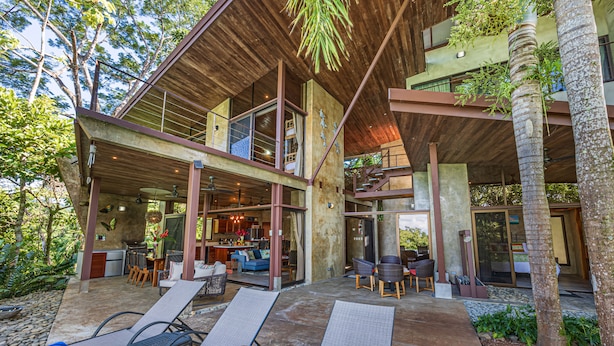 Una casa moderna con un amplio patio, mobiliario exterior y una cocina visible a través de grandes ventanales.