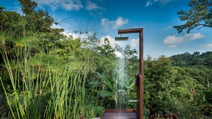 Un sendero de madera que conduce a un área de ducha rodeada de abundante vegetación y un cielo azul despejado.