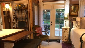 A cozy room with a wooden table, a green upholstered chair, a white bedside table with a lamp, and a bed with floral pillows. - Intercostal Waterfront Private Cottage Retreat Close to Mayo Clinic (Jacksonville)