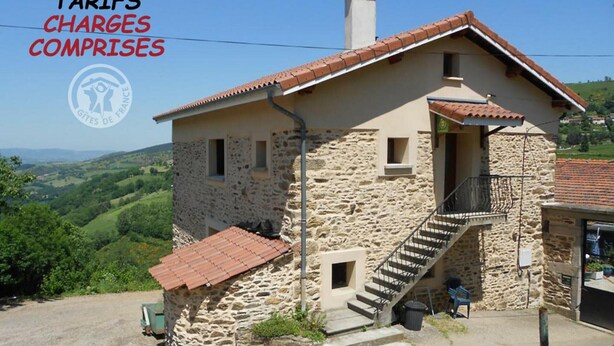A stone house with a red-tiled roof, a staircase leading to a balcony, and a view of the countryside.