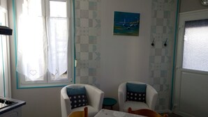 A room with two white armchairs, a small table, a painting of a seascape, and a window with curtains.