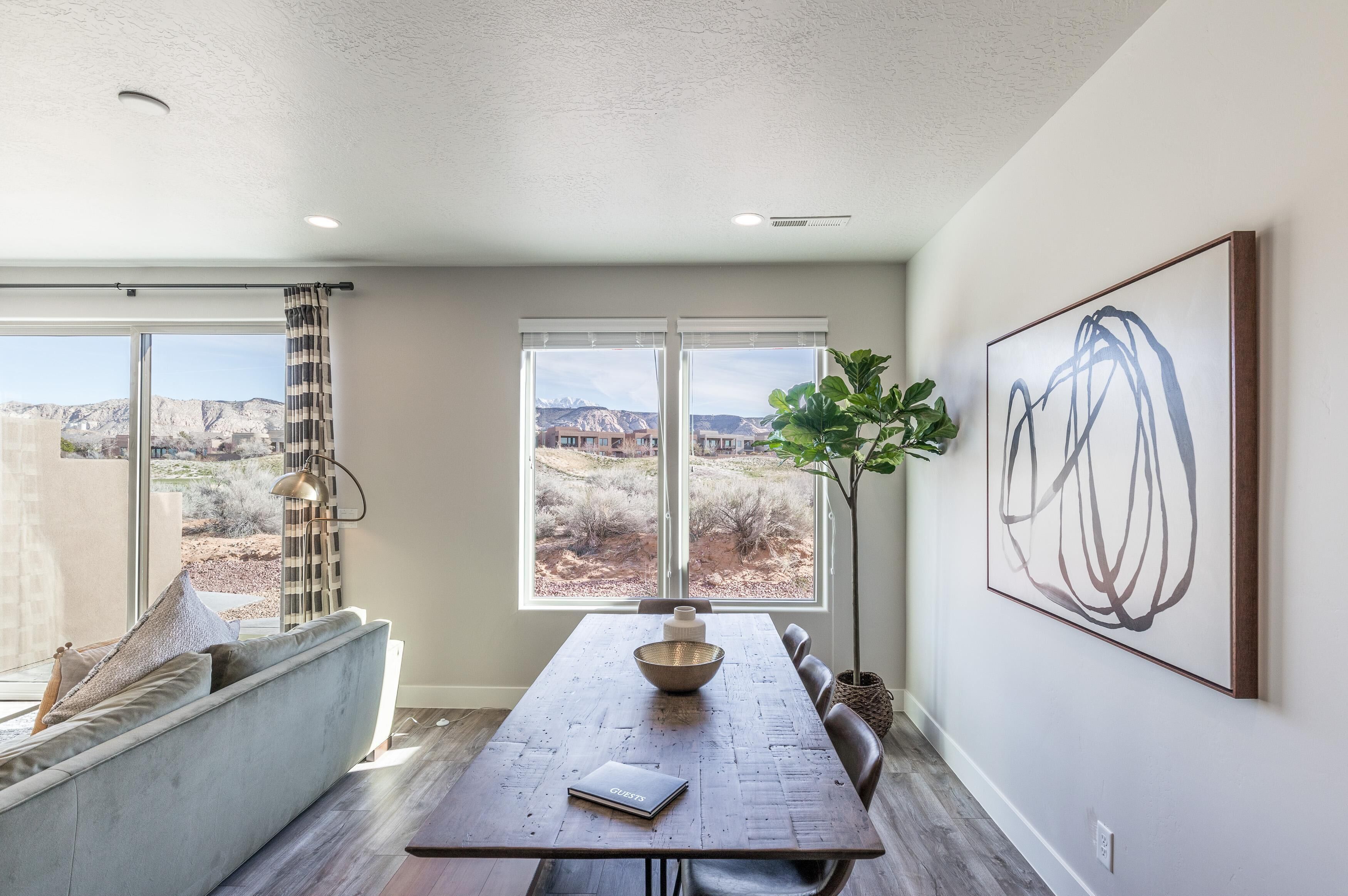 A dining area with a long table, a sofa, and a view of a mountainous landscape through large windows.