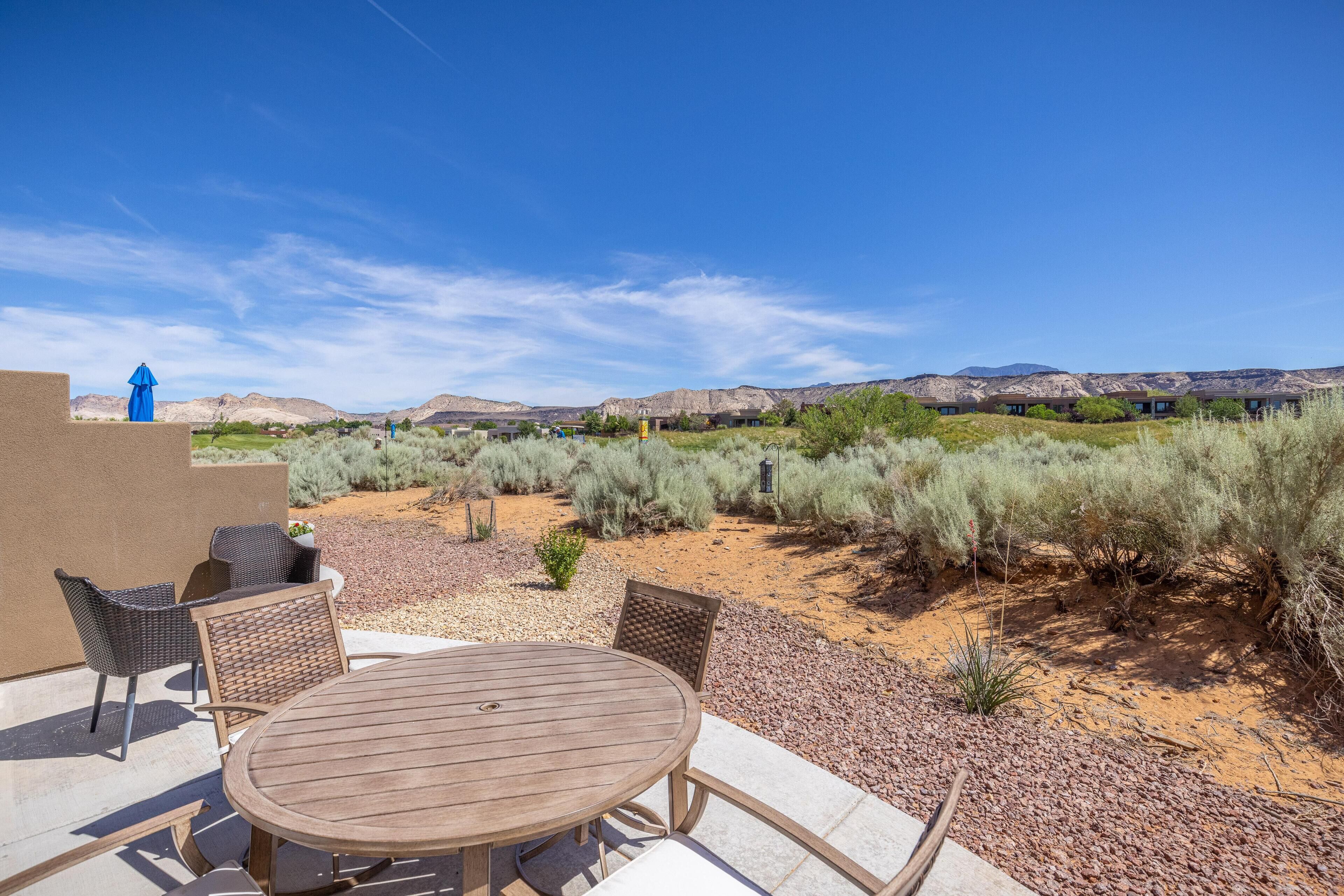 An outdoor patio with a round table and chairs, surrounded by desert vegetation and mountains in the background.