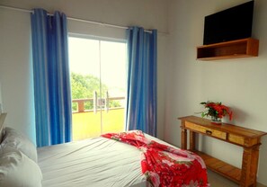 A bedroom with a bed, a wooden nightstand, a TV mounted on the wall, and a balcony with blue curtains. - Pousada Preciosa (Búzios)