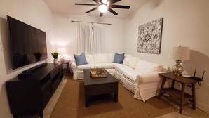A living room with a flat-screen TV, a dark wooden entertainment unit, a white sofa with blue cushions, a coffee table, a patterned tapestry, and a ceiling fan.