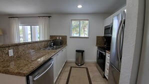 A compact kitchen with granite countertops, stainless steel appliances, a window with blinds, and a trash can.
