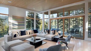 Ein geräumiges Wohnzimmer mit einer großen Eckcouch, einem Couchtisch und einem grauen Sessel. Große Fenster bieten Blick auf Bäume und Berge.