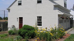 A white two-story house with a red door, a small front porch, and a garden with yellow flowers.