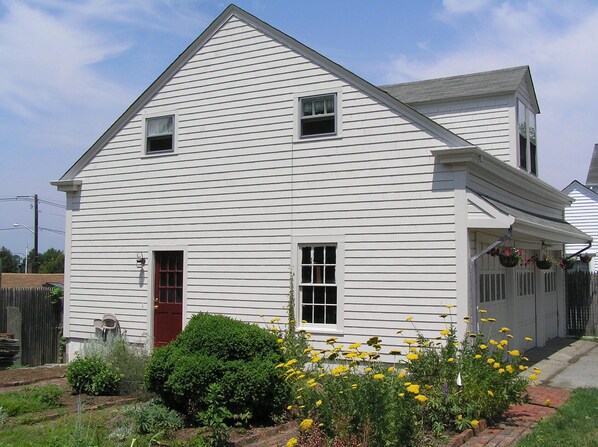 A white two-story house with a red door, a small front porch, and a garden with yellow flowers.