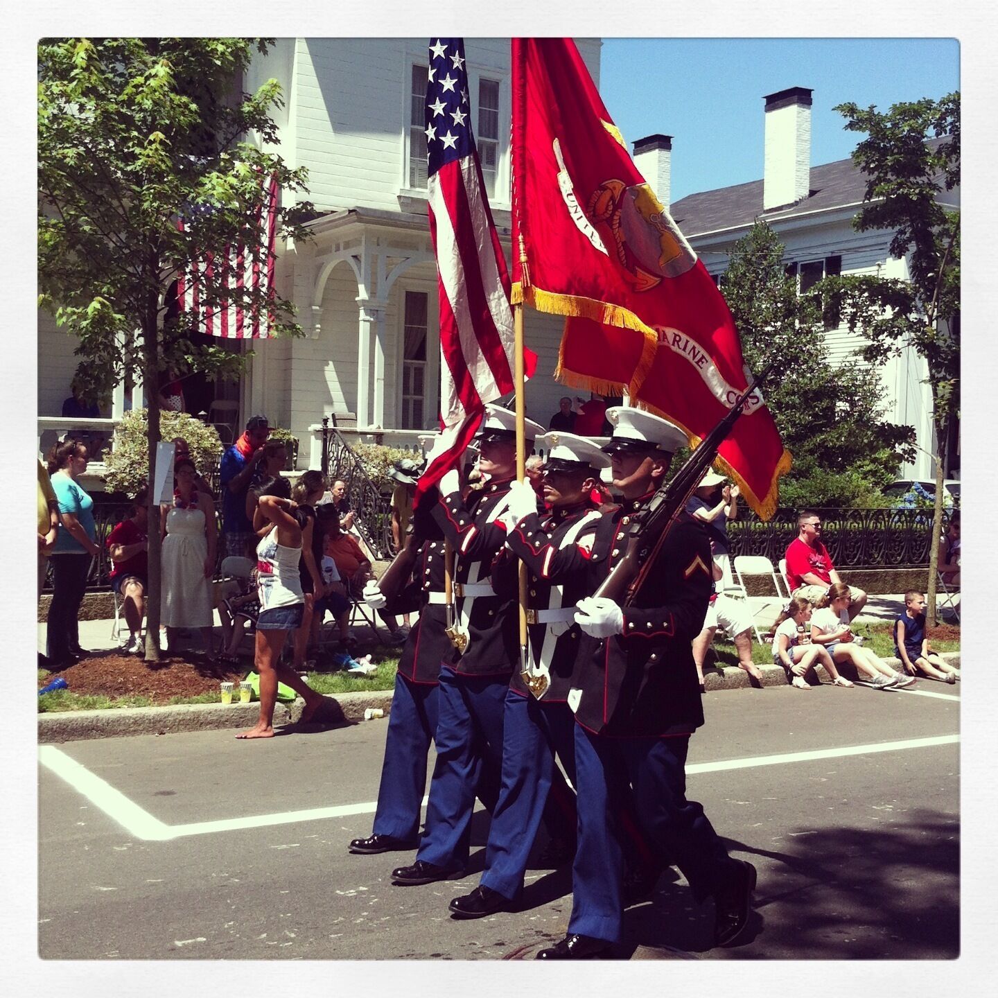 A group of uniformed personnel marching with flags, including an American flag and a Marine Corps flag, in a parade setting.