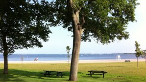 A grassy park area with picnic tables and a bench, overlooking a beach with boats and a clear sky.
