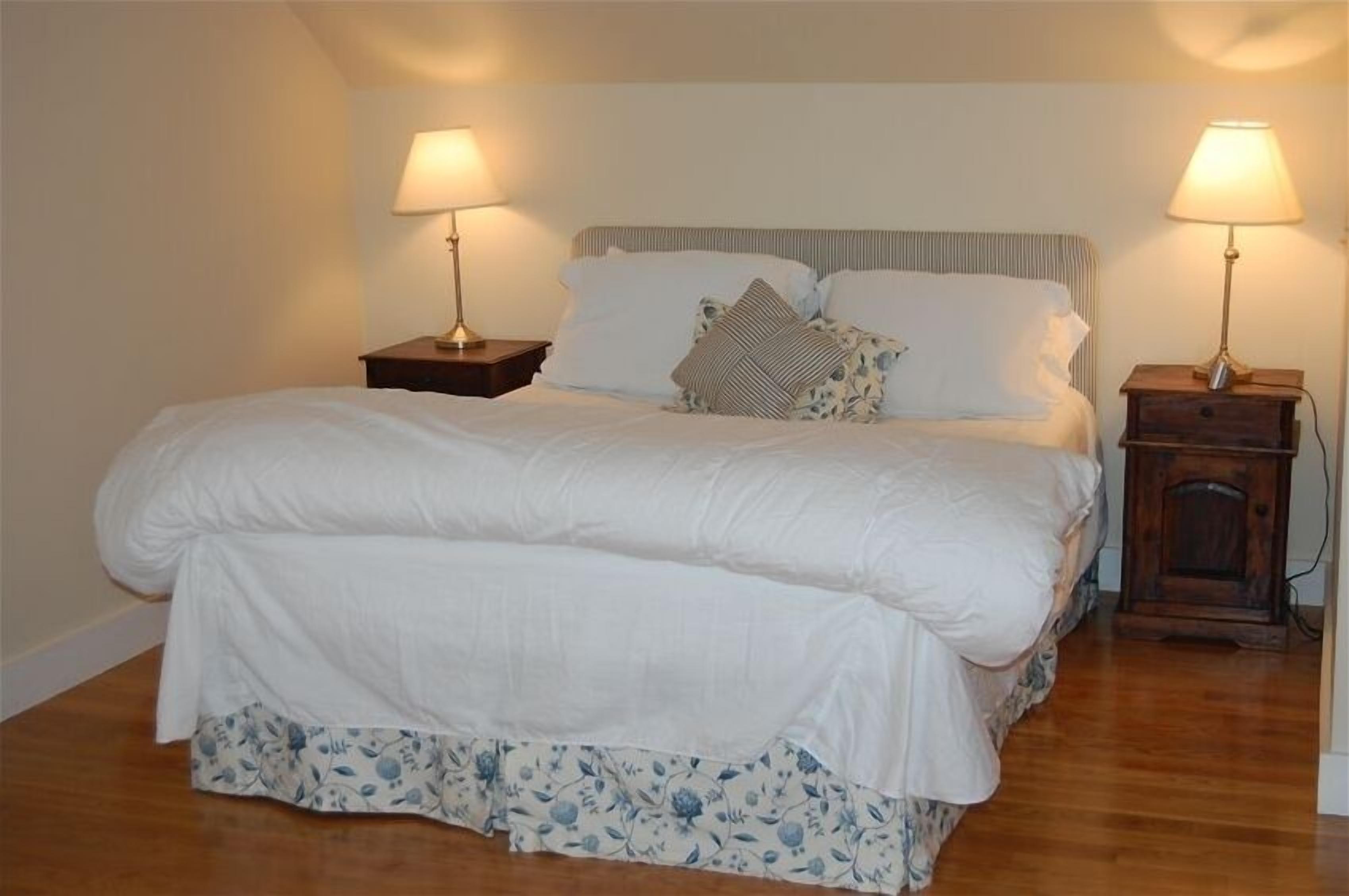 A neatly made bed with white bedding and a floral skirt, flanked by two bedside tables with lamps, in a room with wooden flooring and plain walls.