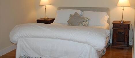 A neatly made bed with white bedding and a floral skirt, flanked by two bedside tables with lamps, in a room with wooden flooring and plain walls.