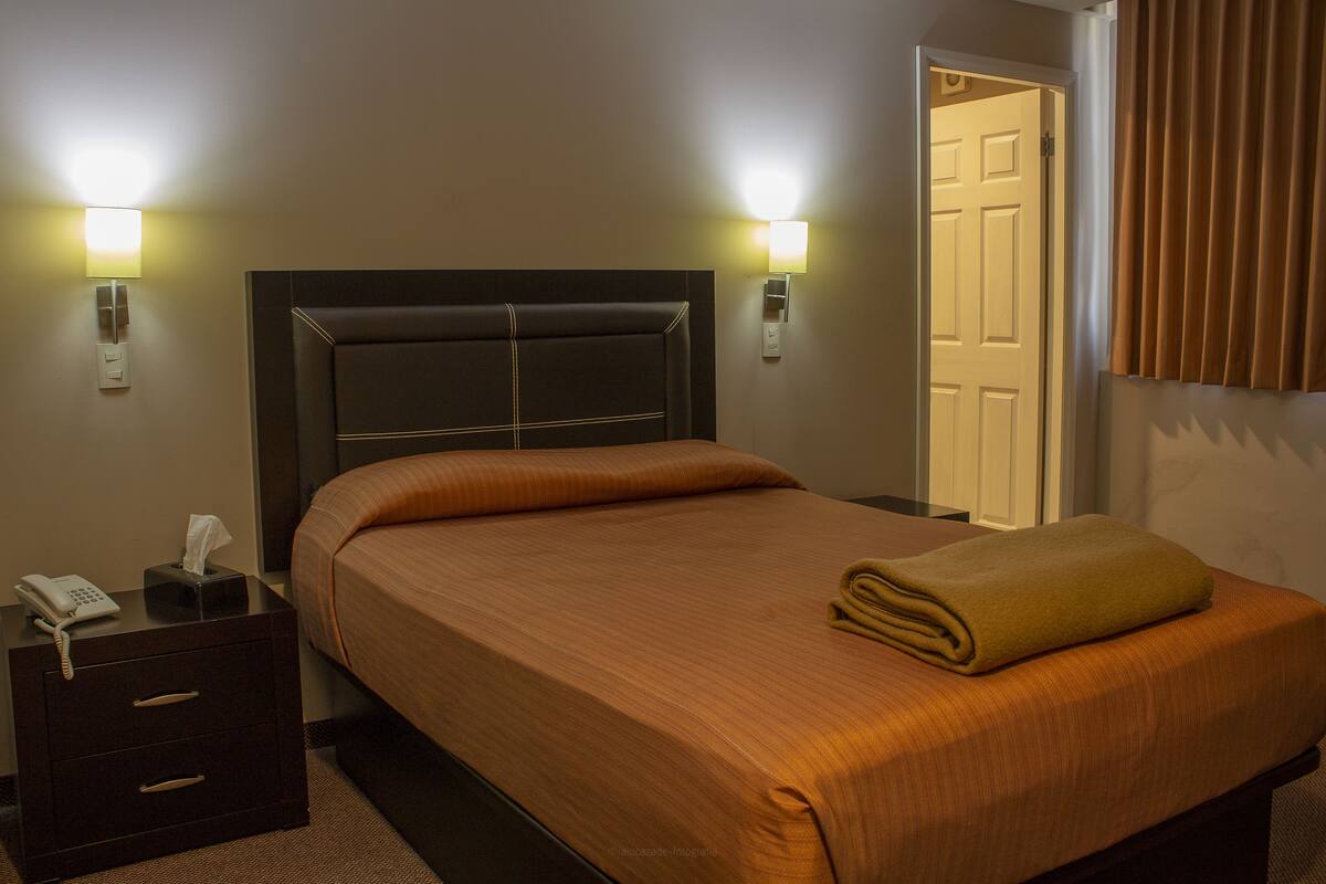 Una habitació d'hotel amb un llit ben fet, làmpades al costat del llit, un telèfon i una porta a la dreta.
