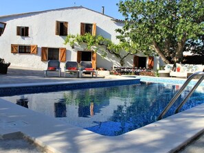 Une piscine avec un bâtiment blanc en arrière-plan, un arbre et du mobilier d’extérieur.