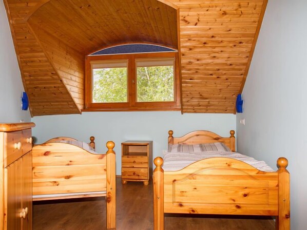 Une chambre avec un plafond en bois incliné, deux lits en bois, une petite table de chevet en bois et une fenêtre donnant sur les arbres.