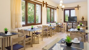 A hotel room with multiple dining tables, chairs, potted plants, a television, and large windows with curtains. - Stella Bay (Samos)