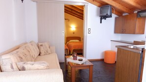 A compact room with a wooden ceiling, a sofa, a small table with a decorative item, a bed with an orange blanket, and a kitchenette with wooden cabinets. - Stella Bay (Samos)