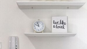 A white wall with two shelves. The top shelf holds three white candles and a black tag. The bottom shelf has a white clock and a sign that says "live like a local". - 8Adriatico2 Contemporary Luxurious Studio (Manila)