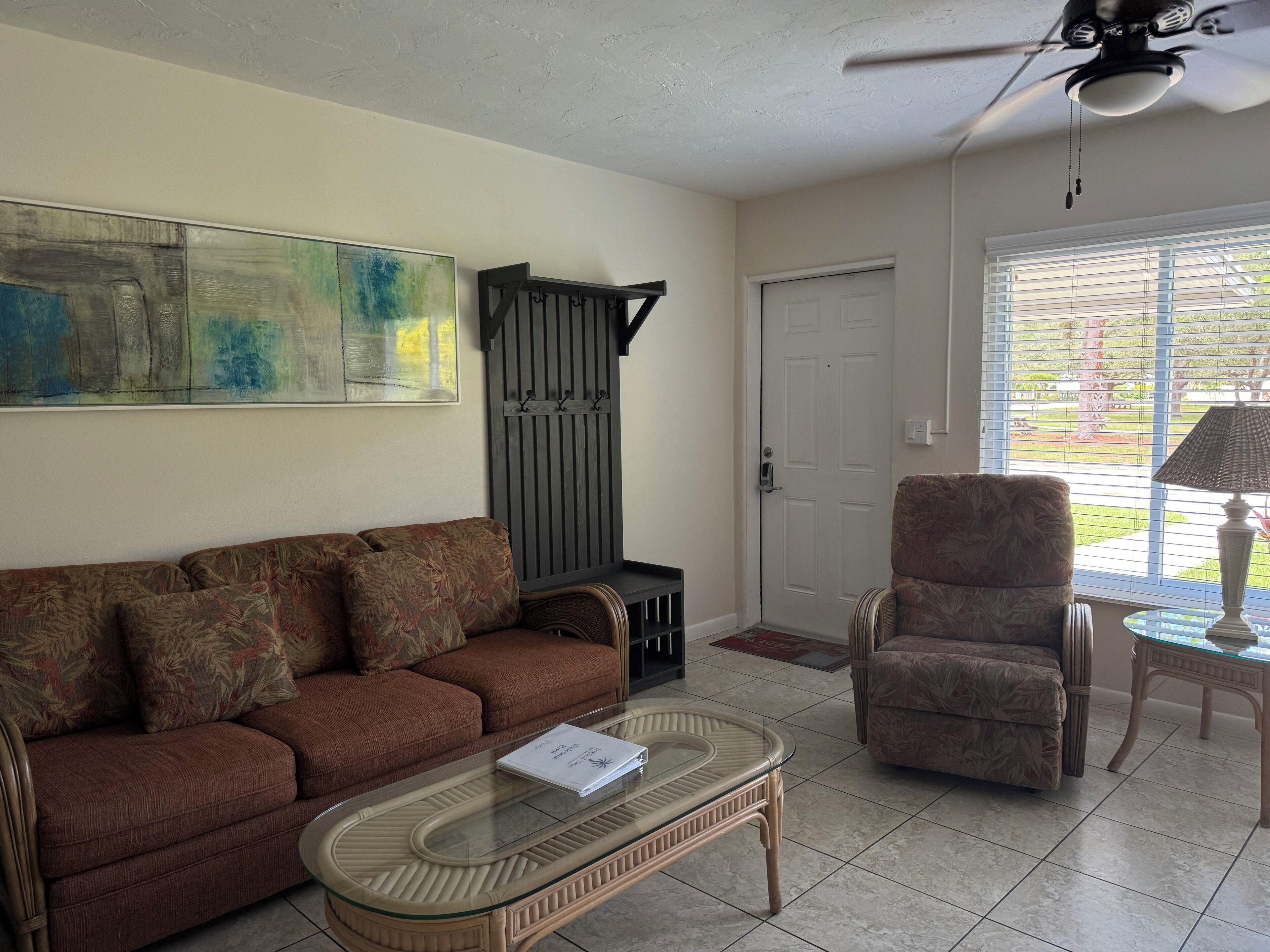 A living room with a brown sofa, a glass coffee table, a patterned armchair, a black entertainment unit, and a ceiling fan.