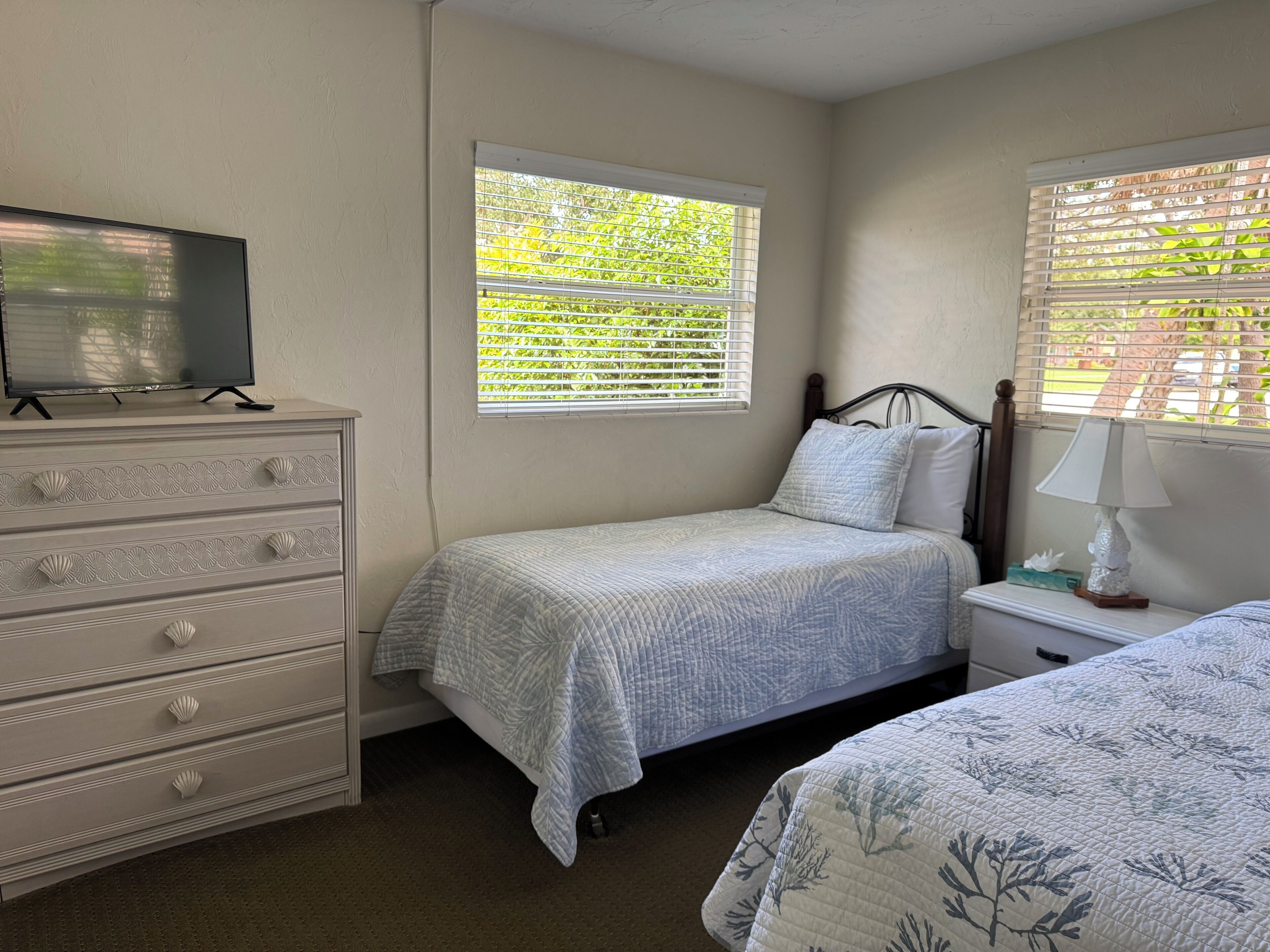 A bedroom with a bed, a dresser, a nightstand, a lamp, and a window with blinds.
