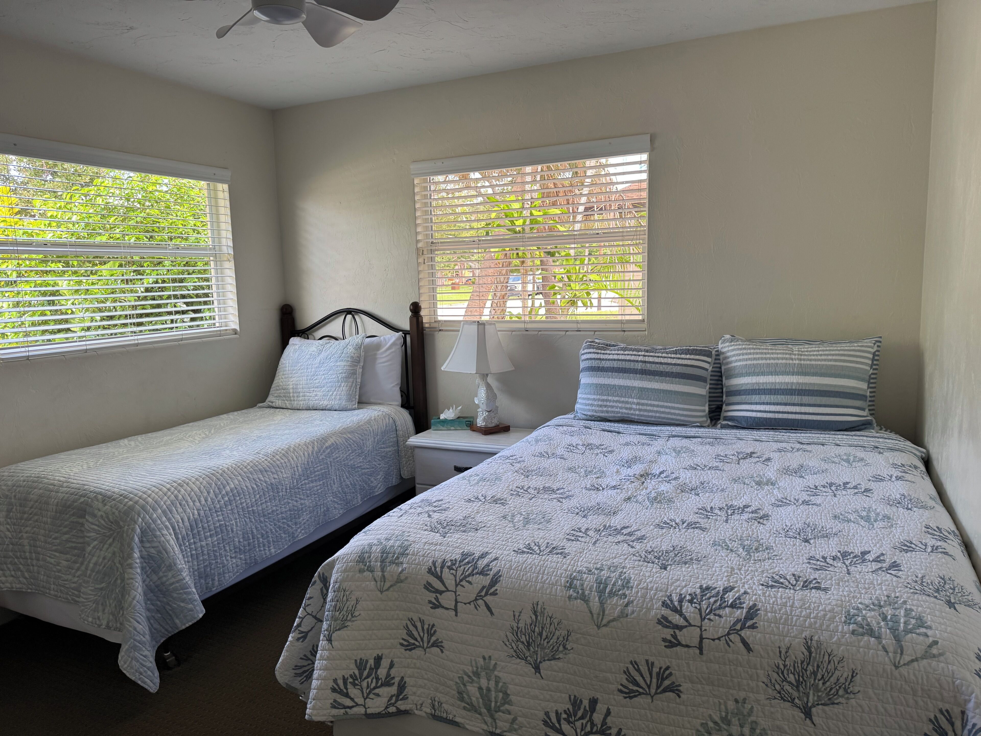 A bedroom with two beds, a nightstand with a lamp, and windows with blinds.