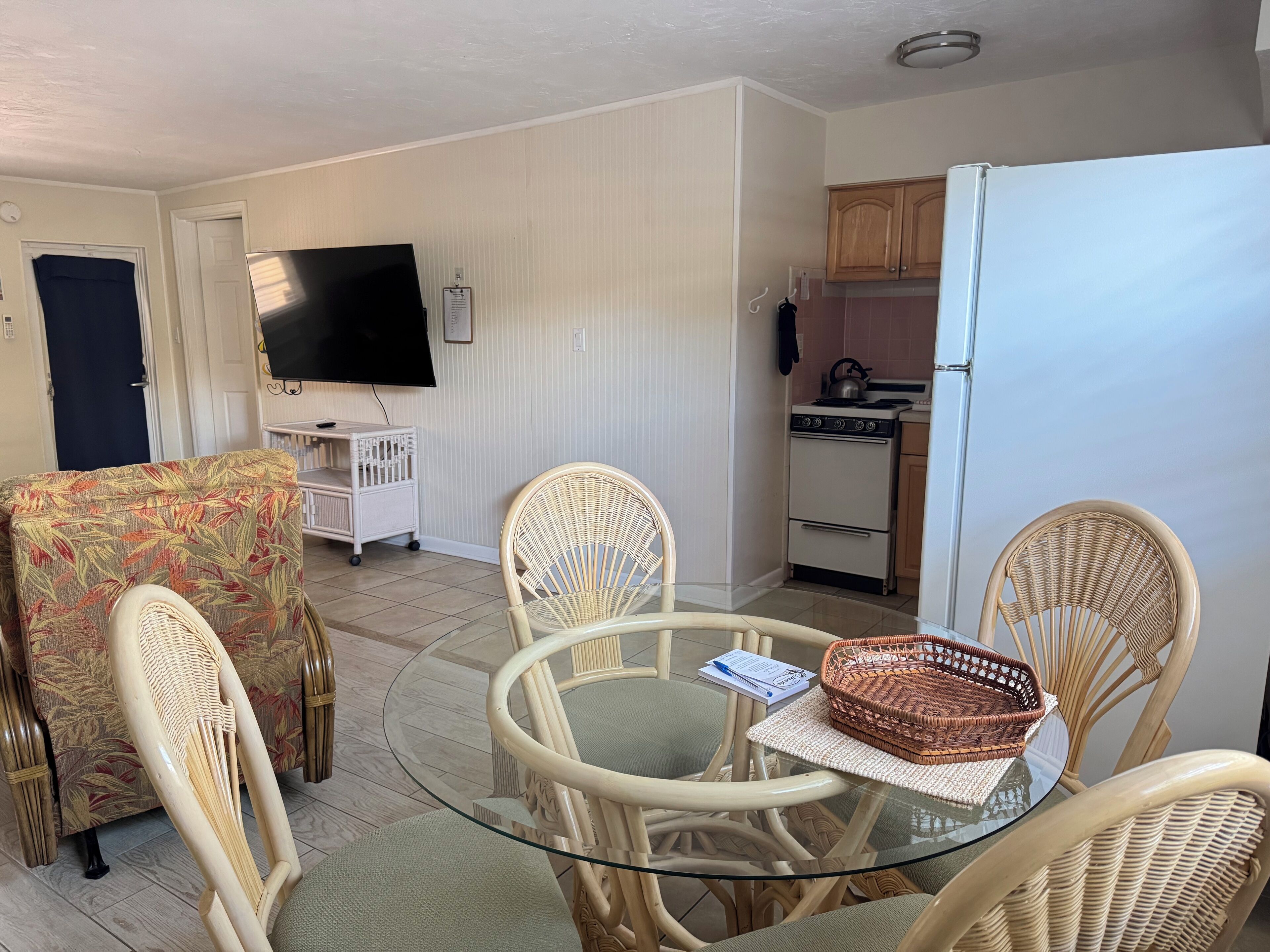 A compact living space with a round glass table surrounded by wicker chairs, a television, a kitchenette, and a door leading to another room.