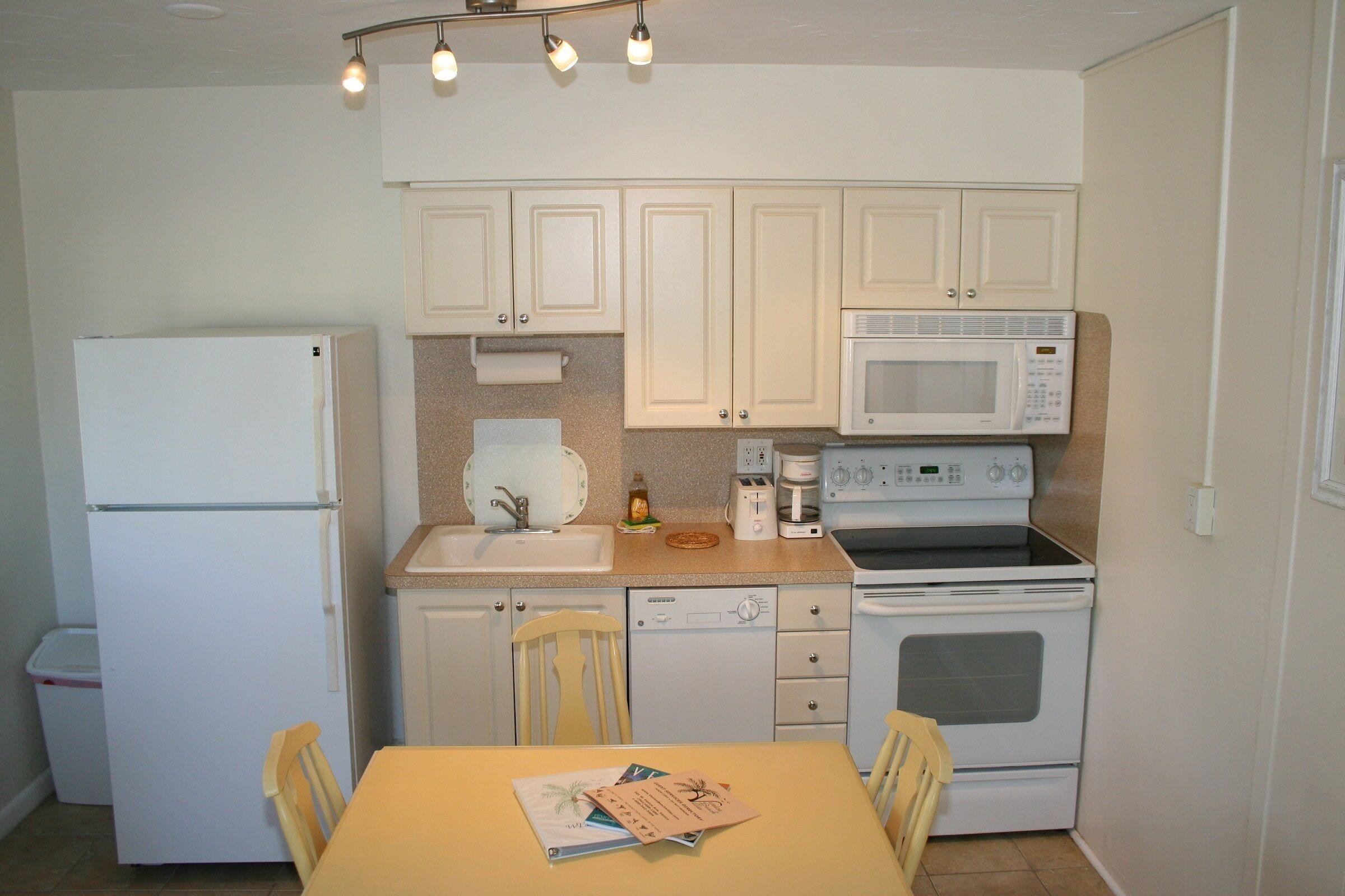 A compact kitchen with white cabinets, a refrigerator, a microwave, and an oven. There is a sink with a faucet, a dish rack, and a table with chairs.