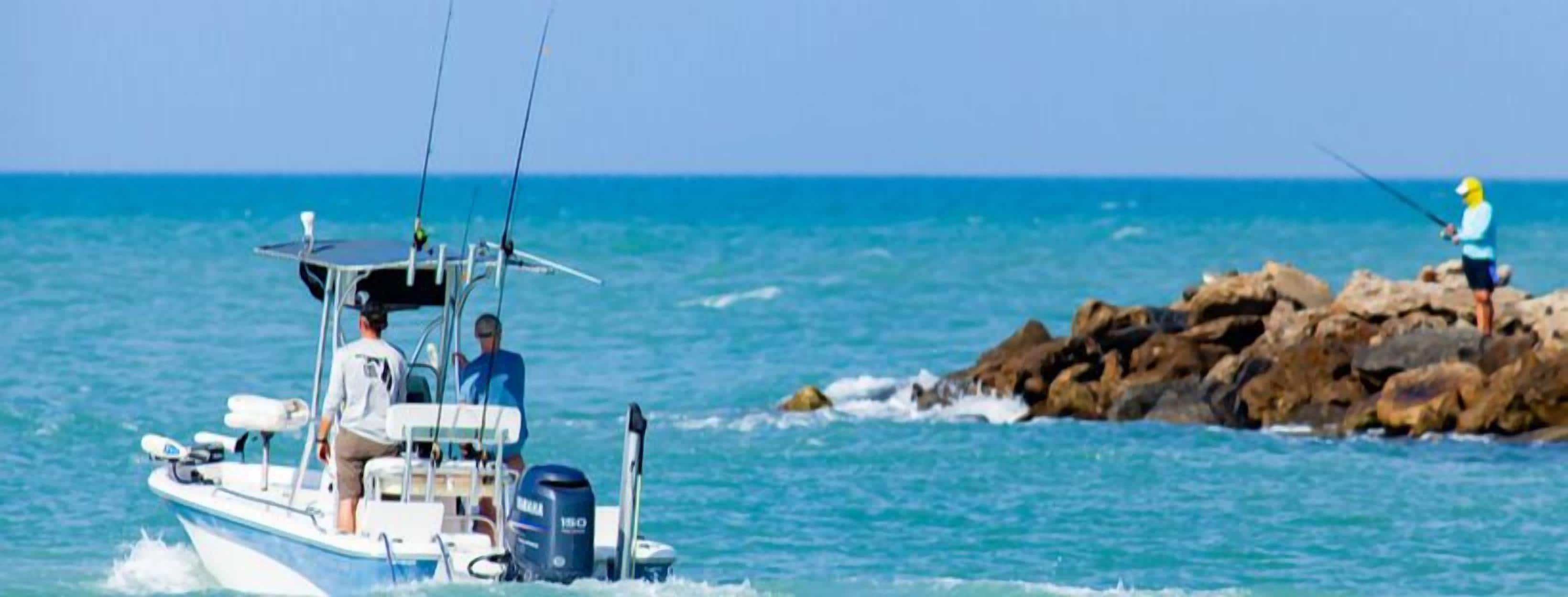 Un bote con dos personas a bordo, una orilla rocosa donde una persona pesca, y agua azul tranquila.