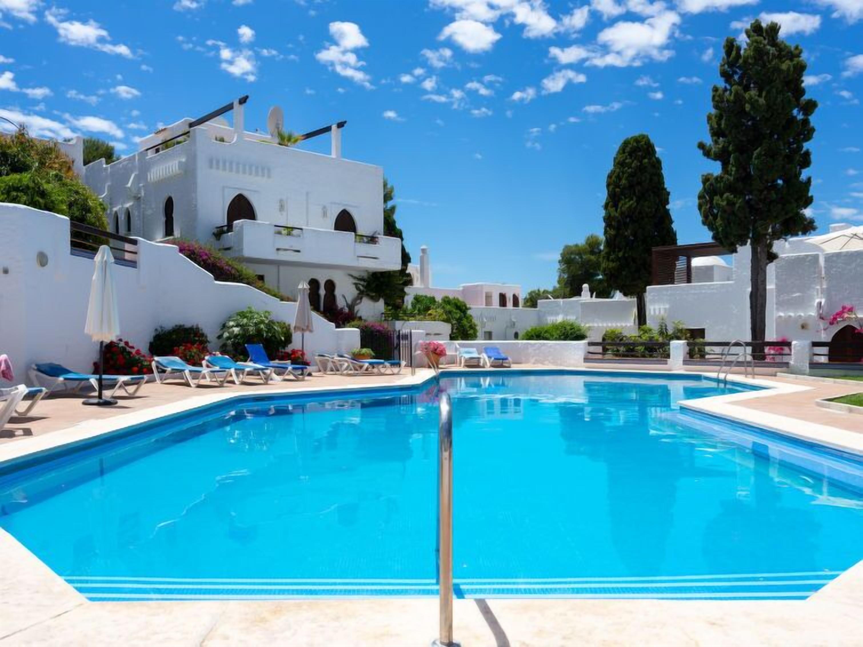 A swimming pool with lounge chairs and umbrellas, surrounded by white buildings with arched windows and balconies.
