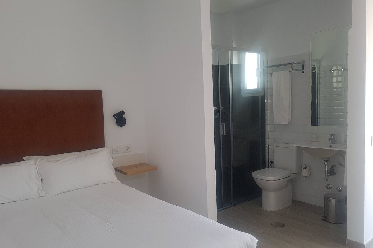 A hotel room with a bed, a small bedside table, a shower, a toilet, and a sink.
