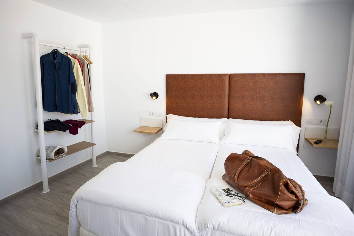 A neatly made bed with white linens, a brown leather bag on the bed, and a clothing rack with various garments.