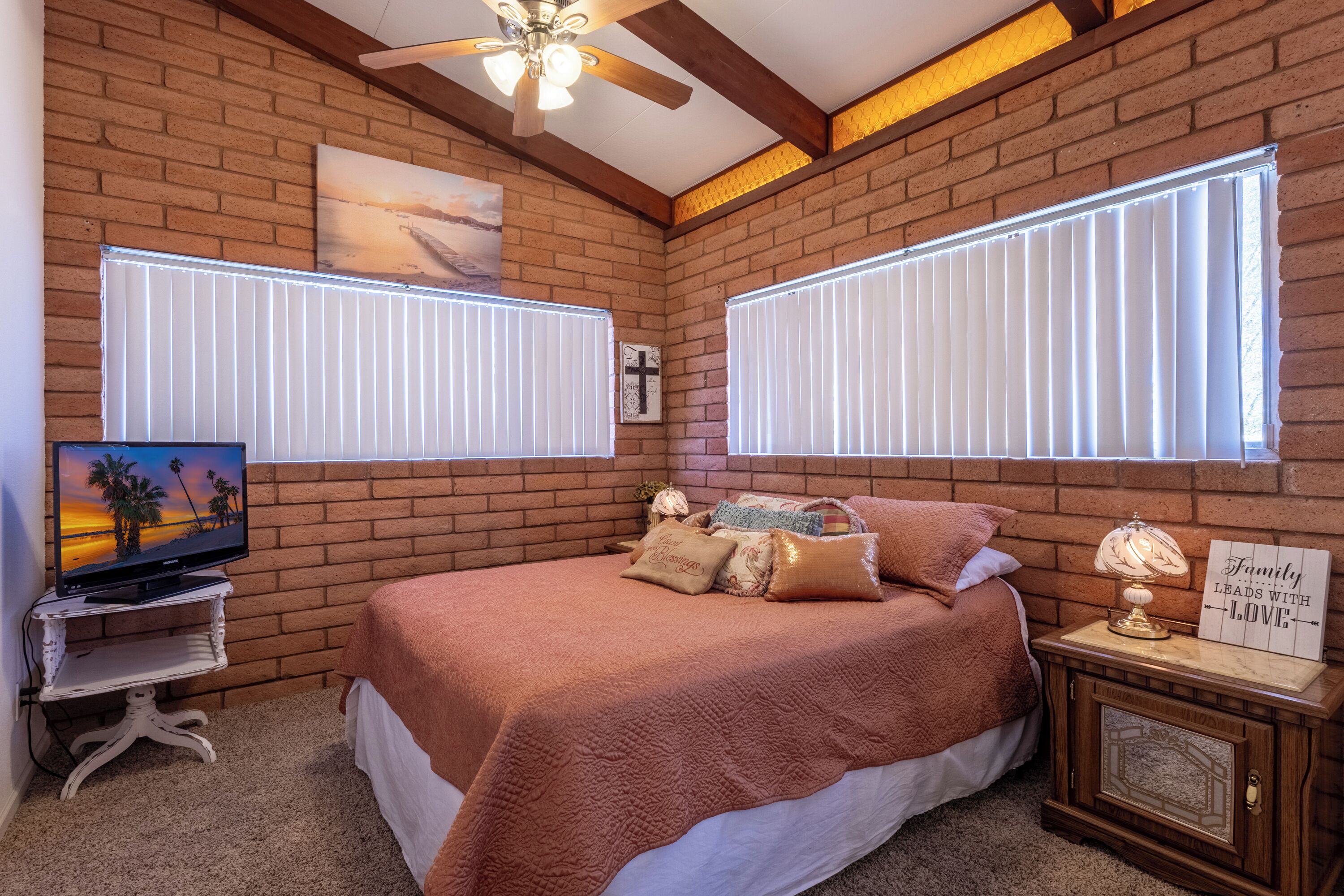 A bedroom with a brick wall, a bed with a pink blanket, a television on a white stand, and a nightstand with a lamp and a sign.