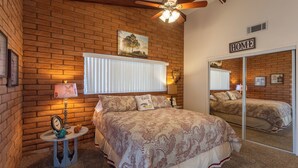 A bedroom with a brick wall, a ceiling fan, a bed with a patterned bedspread, a bedside table with a lamp, and a mirror. - Upgraded lake view (Lake Havasu City)