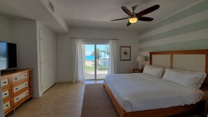 Ein Hotelzimmer mit Bett, Fernseher, Deckenventilator, Nachttisch mit Lampe und Blick auf den Ozean durch eine Schiebetür.