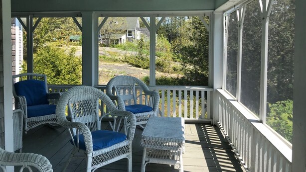 A covered porch with white wicker chairs and blue cushions, overlooking a residential area with trees and houses.