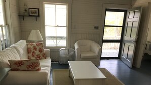 A living room with a white sofa, a white table, a chair, a floor fan, and a window with blinds.